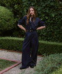 A woman stands in a garden with lush green bushes, wearing the Denim Newport Trouser and a dark blue denim shirt tucked in with a black belt. Her long hair is down and her hands are on her hips.
