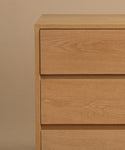 The Dune Dresser features a simple, modern design with clean lines and three visible drawers, crafted in white oak style and set against a beige background.
