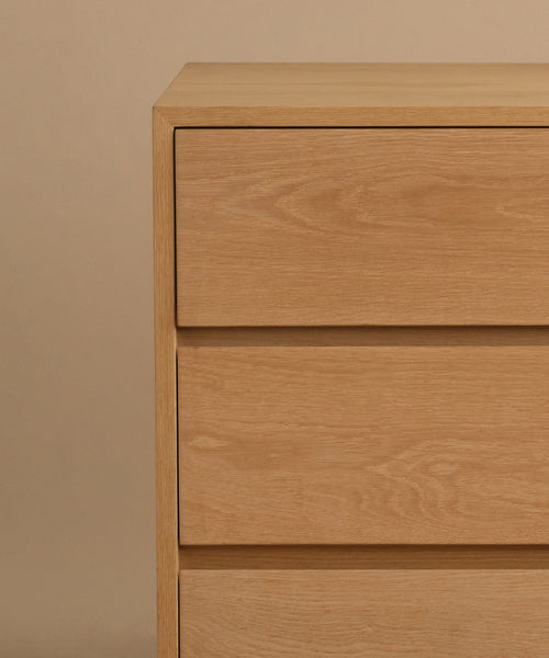 The Dune Dresser features a simple, modern design with clean lines and three visible drawers, crafted in white oak style and set against a beige background.