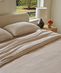 A neatly made bed with the Dusk Linen Duvet Cover sits beside a wooden nightstand topped with a black lamp and a vase of flowers. Sunlight pours through the window above, illuminating lush greenery outside.