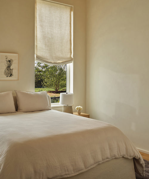 A minimalist bedroom with a neatly made bed featuring the Dusk Linen Duvet Cover, light fabric shades on the window, a small bedside table topped with a lamp and vase, and a framed drawing on the cream-colored wall.