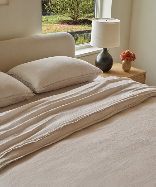A neatly made bed with the Dusk Linen Duvet Cover sits beside a wooden nightstand topped with a black lamp and a vase of flowers. Sunlight pours through the window above, illuminating lush greenery outside.