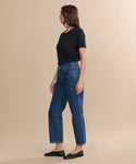 A woman in profile stands against a plain beige background, wearing a black short-sleeve shirt, blue Easy Straight Crop jeans with a relaxed leg, and black flats.