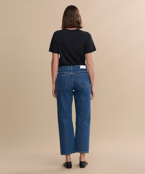 A person with straight brown hair faces away, wearing a black t-shirt, black shoes, and blue RE/DONE Easy Straight Crop jeans with a relaxed leg, against a plain beige background.