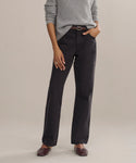 A person wears the Easy Straight Crop luxury jeans in a straight cropped leg and mid-rise fit, paired with a light gray sweater, brown belt, and brown shoes, standing with one hand in their pocket against a plain background.