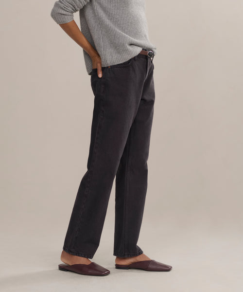A person stands sideways with one hand on their hip, wearing a gray sweater and the Easy Straight Crop luxury jeans in a mid rise, straight cropped fit, paired with brown flat shoes against a neutral background. Their face is not visible.