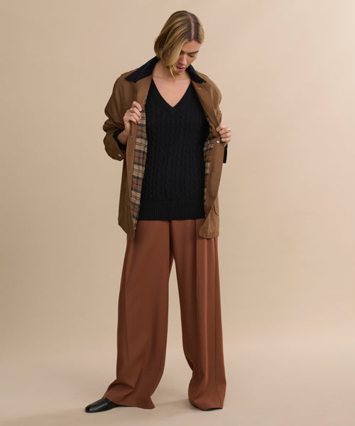 A person stands against a beige background, looking down and holding open the Eaton Jacket—a garment-dyed canvas chore coat with plaid lining—worn over wide-legged brown pants and a black V-neck sweater.