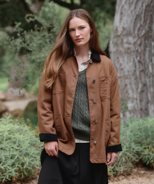 A woman with long brown hair stands outdoors in the Eaton Jacket—a brown garment-dyed canvas chore coat—worn over a green sweater and striped shirt, with greenery and a large tree trunk in the background.