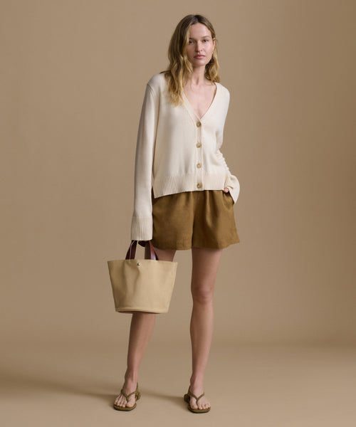 Woman wearing an ivory cardigan with brown shorts carrying a handbag against a beige wall. 