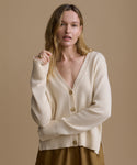 Woman wearing an ivory cardigan with brown shorts against a beige wall. 