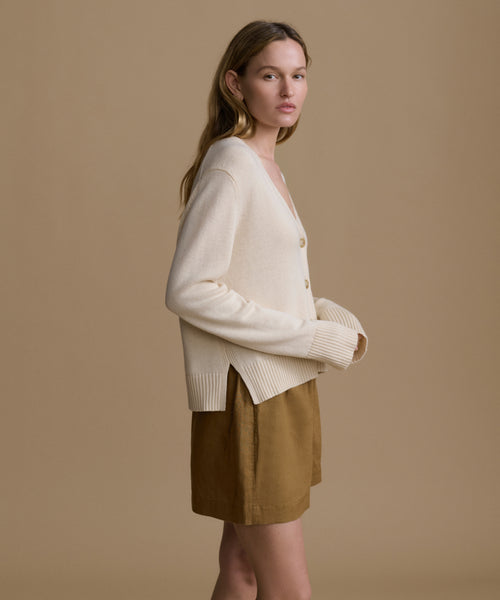 Woman wearing an ivory cardigan with brown shorts against a beige wall. 