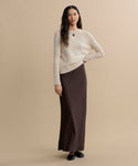 A woman with long wavy hair stands against a beige background, wearing the Everyday Sweater in cream, paired with a long brown skirt and black shoes. Her arms are relaxed at her sides and she has a calm expression.