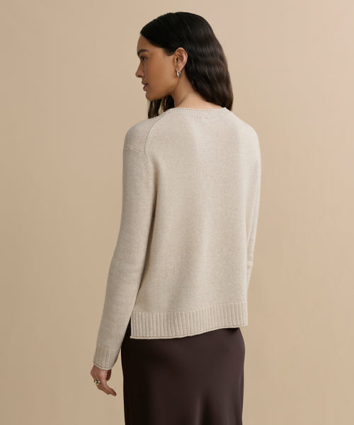 A woman with long dark hair, wearing the Everyday Sweater and a dark brown skirt, stands turned slightly to the side against a plain beige background.