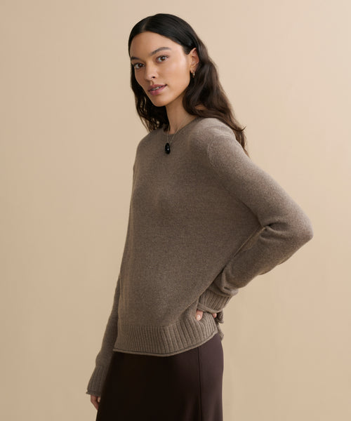A woman with long dark hair wears the Everyday Sweater in lightweight brown knit with a matching skirt, standing sideways against a beige background and looking at the camera with a neutral expression.