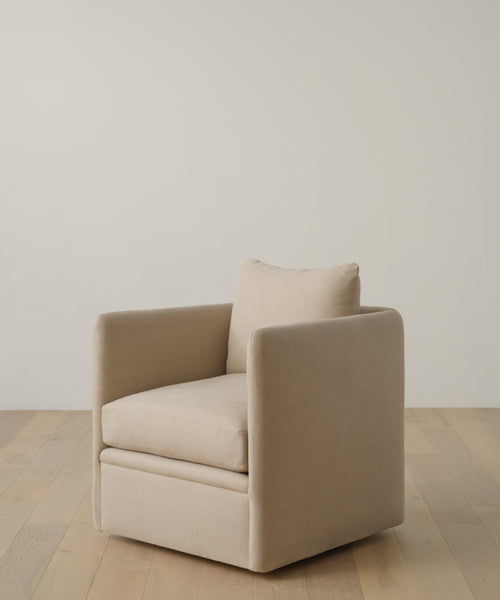 A modern armchair upholstered in Fabric Swatch Sand Hemp with a boxy silhouette, loose back cushion, and swivel base rests on a light wooden floor by an off-white wall—an ideal match for the Dume Sofa.