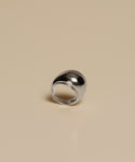 The Felix Dome Ring, a shiny silver dome-shaped white gold piece, rests on a plain beige surface and reflects light with its smooth, polished finish.