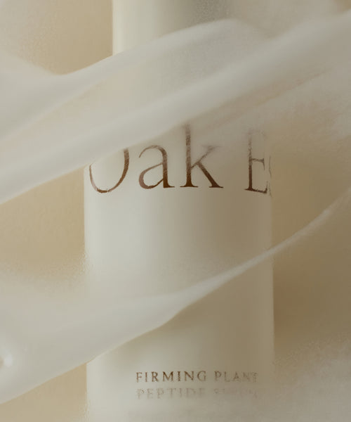 A frosted white bottle, draped in sheer fabric, displays “Firming Plant Peptide Serum,” suggesting an anti-aging and collagen-boosting formula set against a soft, neutral background.