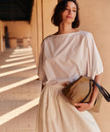 Woman wearing a white blouse, ivory skirt and brown bag against a wall outside. 