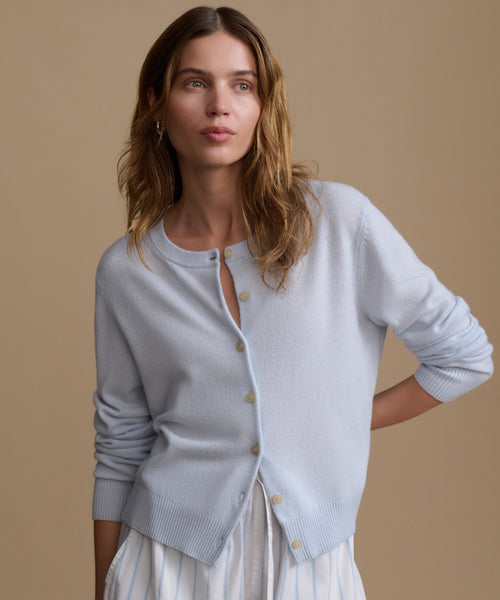 Woman wearing a light blue cardigan with blue and white stripe shorts against a beige wall.