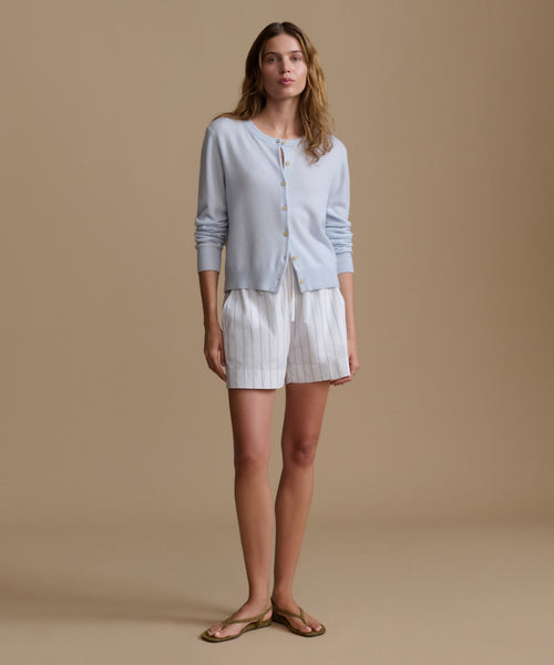 Woman wearing a light blue cardigan with blue and white stripe shorts against a beige wall.