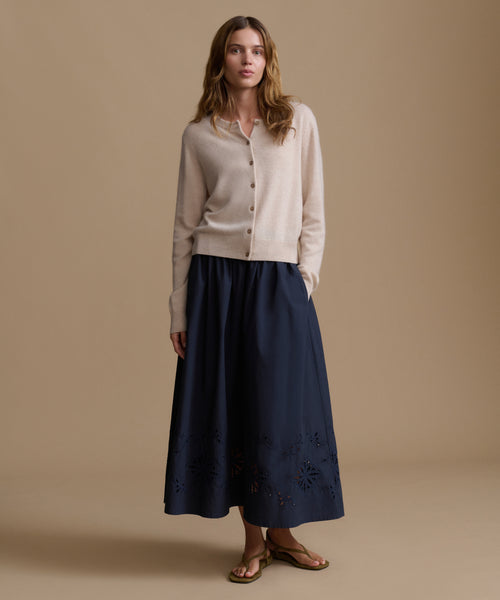 Woman wearing an oat cashmere cardigan with a blue skirt against a beige wall. 