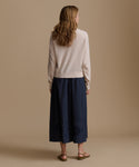 Woman wearing an oat cashmere cardigan with a blue skirt against a beige wall. 