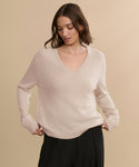 A woman with shoulder-length brown hair wears the Flynn Cashmere Sweater in light beige and black pants, standing against a plain beige background while looking down to her left.