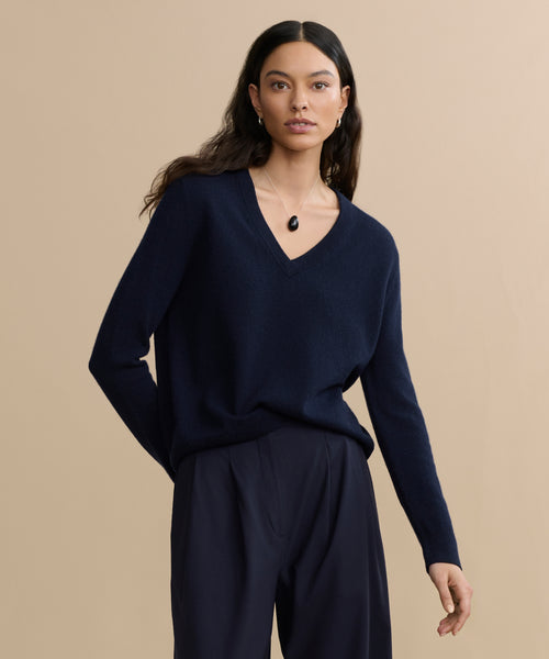 A woman with long dark hair wears the Flynn Cashmere Sweater, a relaxed fit navy V-neck knit, paired with loose dark trousers and a black pendant necklace, standing against a plain beige background.