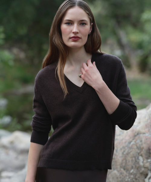 A woman with long brown hair stands outdoors amid rocks and greenery, looking at the camera with a neutral expression and one hand near her collar, wearing the Flynn Cashmere Sweater in a relaxed fit V-neck style.