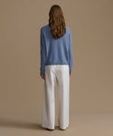 Woman wearing a blue cashmere sweater and white pants against a beige background