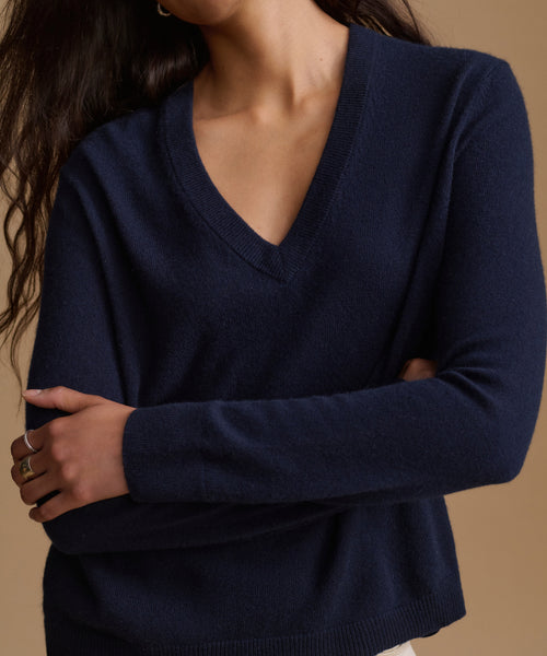 A close-up of a woman wearing a navy blue V-neck long-sleeve knit sweater against a warm beige background.