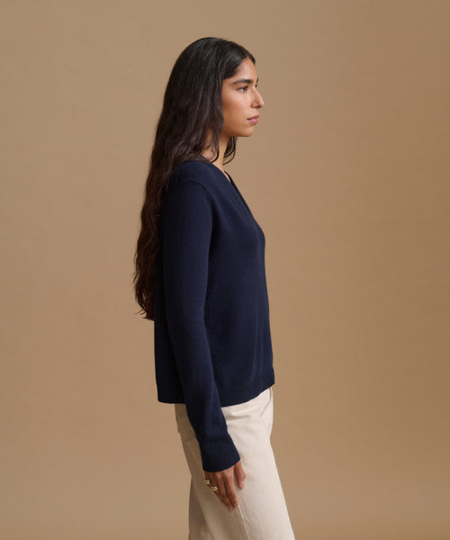 A woman in side profile wearing a navy blue long-sleeve knit sweater and cream-colored pants, standing against a solid warm beige studio background.