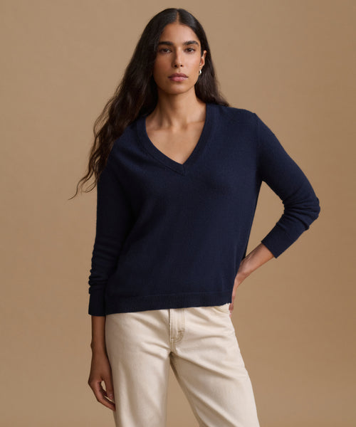 A woman modeling a navy blue V-neck knit sweater with cream pants, standing against a solid beige studio background.