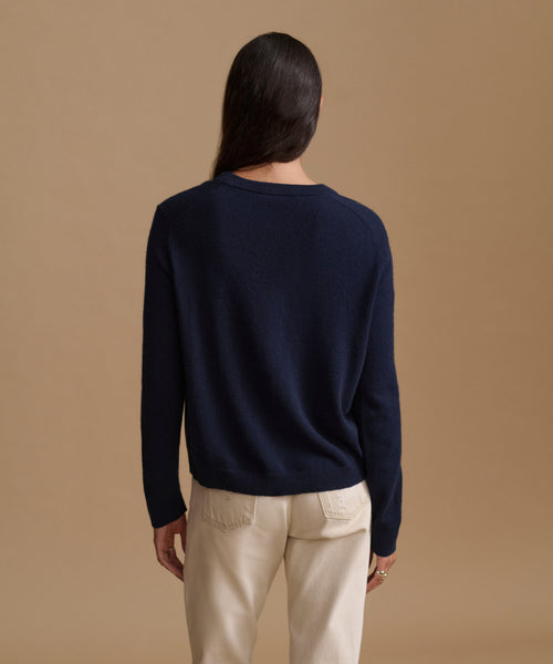 A woman viewed from behind wearing a navy blue long-sleeve knit sweater and cream-colored trousers, standing against a solid tan studio background.