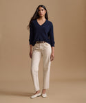 A woman modeling a navy blue V-neck sweater, cream-colored straight-leg jeans, and white flat shoes against a solid beige studio background.
