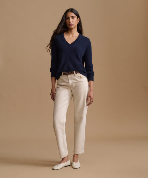A woman modeling a navy blue V-neck sweater, cream-colored straight-leg jeans, and white flat shoes against a solid beige studio background.