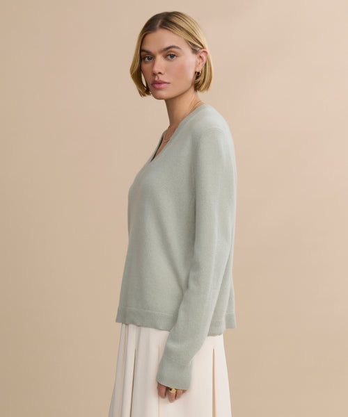 A woman with short blonde hair, wearing the relaxed fit Flynn Cashmere Sweater in light gray and a cream skirt, stands in profile against a beige background.