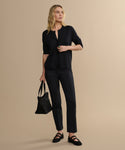 A woman with blonde hair stands against a beige background, wearing the Forme Pant, a black long-sleeve top, black shoes, and holding a black tote bag in her left hand while looking slightly to the side.