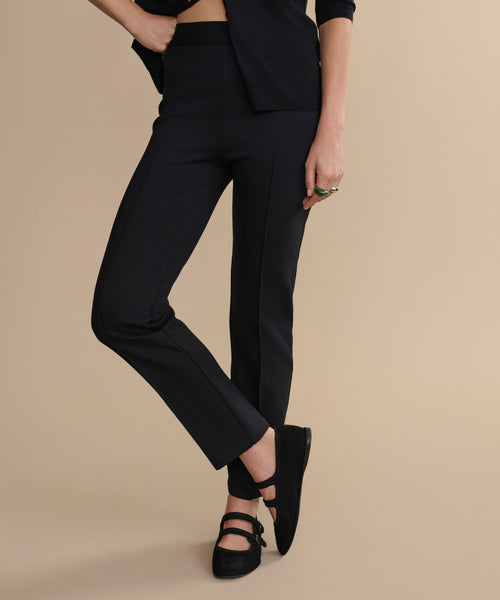 A person models the black Forme Pant with a matching black top and Mary Jane shoes against a beige backdrop. One arm is on their hip, and their face is not shown.