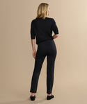 A woman with blonde hair, wearing a black long-sleeve top and the Forme Pant—a black stretch knit style with pintuck detail—stands facing away from the camera against a plain beige background.