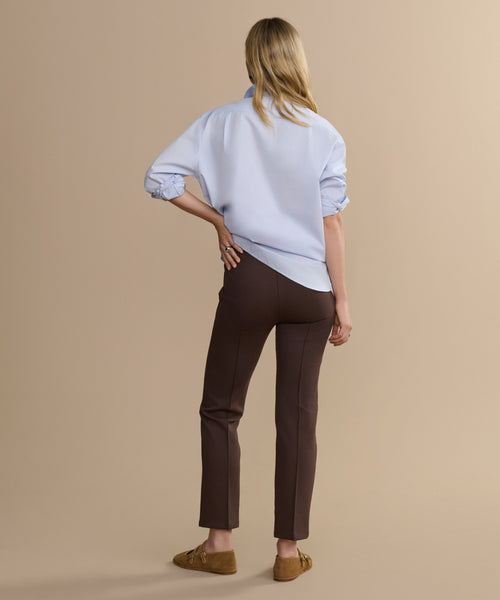 A woman faces away in brown Forme Pants, a light blue collared shirt with rolled-up sleeves, and brown loafers, set against a beige background.