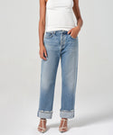 A person in a white sleeveless top, light blue Fran Jean relaxed leg jeans with an untacked folded cuff, and white strappy heeled sandals stands against a plain light background.