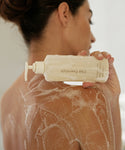 A woman with her back to the camera holds a bottle from the Fresh Start Set. Her shoulder and upper back are covered in a lather of soap as she begins to cleanse and hydrate her skin.