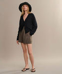 A woman wearing a black wide-brim hat, a black V-neck long-sleeve top, taupe shorts, and the Gia Sandal stands against a plain beige background.