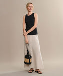 A woman with light hair pulled back stands against a beige background, wearing a sleeveless black top, long beige skirt, woven straw bag with black accents, and the Gia Sandal—an effortless summer sandal—her expression is relaxed.