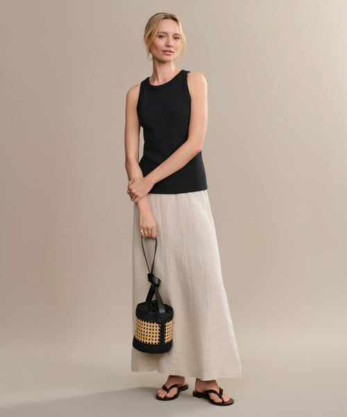 A woman with light hair pulled back stands against a beige background, wearing a sleeveless black top, long beige skirt, woven straw bag with black accents, and the Gia Sandal—an effortless summer sandal—her expression is relaxed.