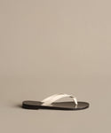 The Gia Sandal features white crisscross straps and a flat black sole, showcased on a light taupe background.