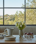 A wooden table set with plates, the Universal Low Glass Set of 4 by Vincent Van Duysen, a ceramic pitcher, a vase of white tulips, and apples sits before a large window overlooking trees and a distant landscape.