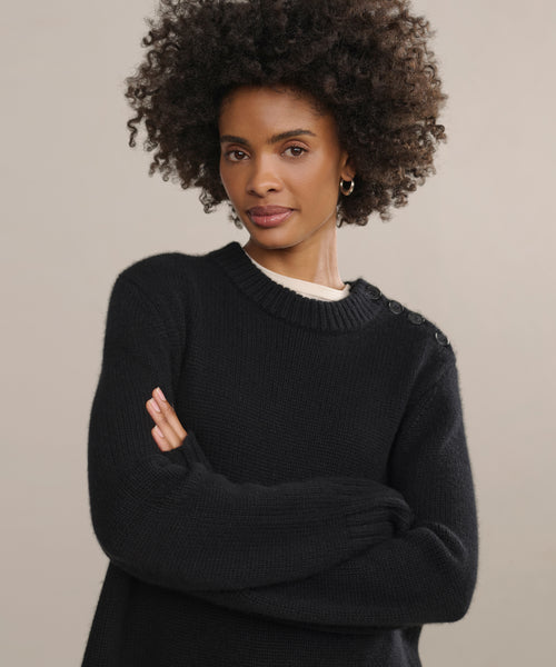 A woman with curly hair stands confidently with arms crossed, wearing the Grace Crewneck—a black cashmere sweater with buttoned shoulders—against a neutral background, effortlessly highlighting timeless luxury knitwear.