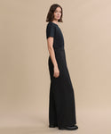 A woman stands sideways against a beige background, wearing a black short-sleeve top, the Greta Trouser—high-waisted, wide-leg tailored trousers in black with a relaxed drape—and black shoes.
